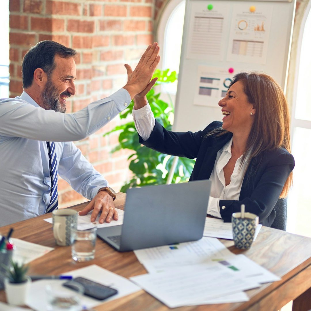 Sales Consultancy Two colleagues joyfully high-fiving at a desk with a laptop and documents.